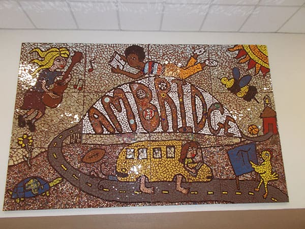 ceramic tile mosaic mural featuring whimsical imagery of students, a school bus, and the ambridge bridge