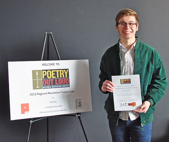 2014-2015 regional Poetry Out Loud winner Drew Praskovich, smiling, holding a winner's certificate and standing by a sign for Poetry Out Loud
